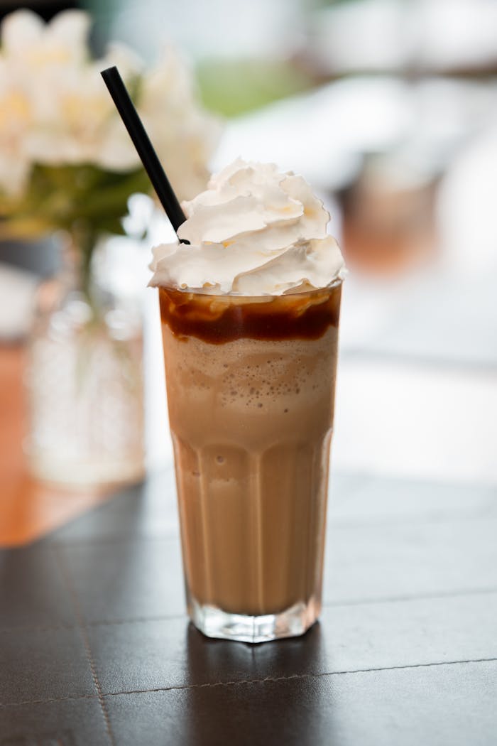 Tall glass of iced coffee topped with whipped cream and caramel sauce, featuring a straw in a cafe setting.